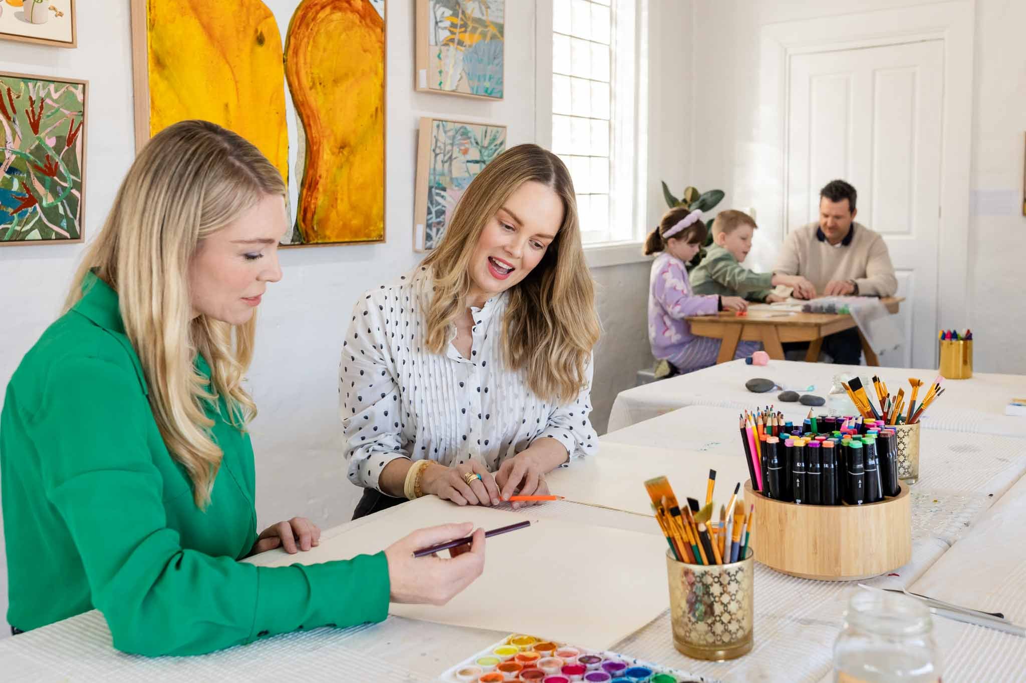 People engaging in arts and crafts activity at a table, with colorful art supplies including markers, colored pencils, and paints, in a room decorated with vibrant artwork.