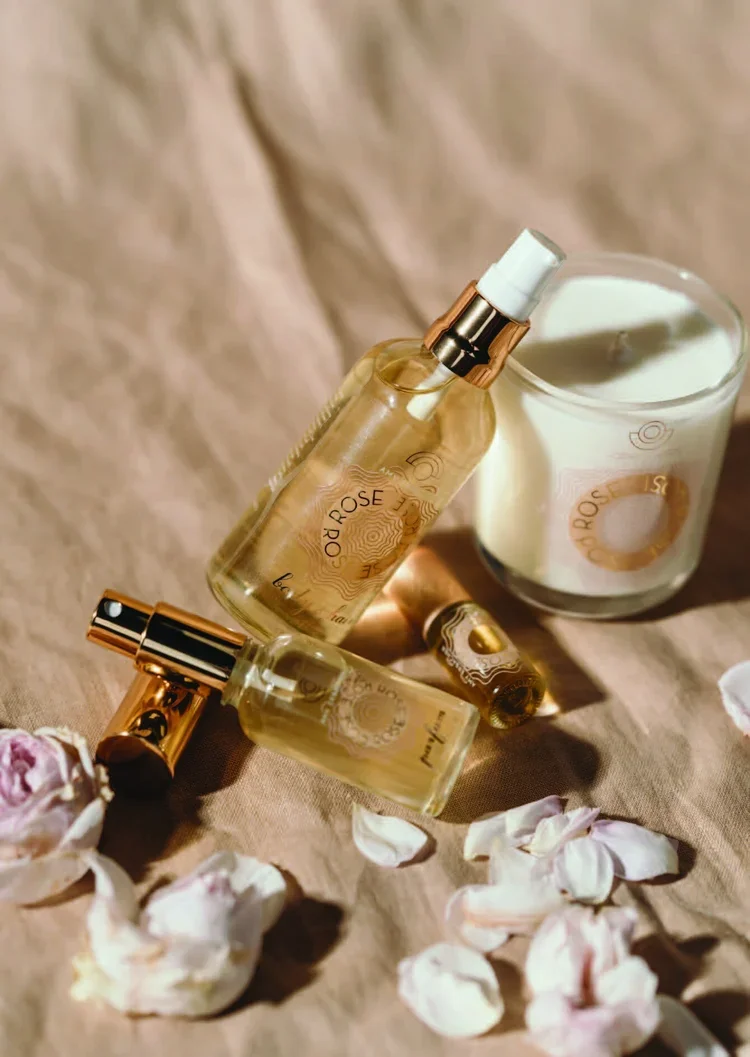 Scarlet Rose perfume bottles and a lit candle on a beige fabric surface, surrounded by white rose petals.