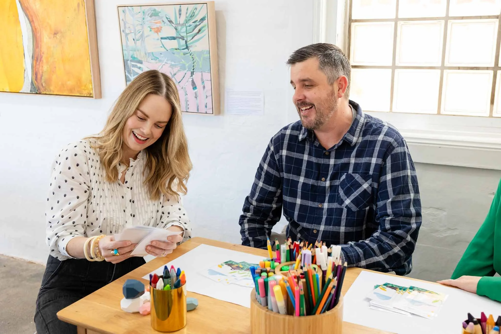 Two people smiling and looking at a piece of paper at a table with colorful art supplies in a bright art studio or classroom.