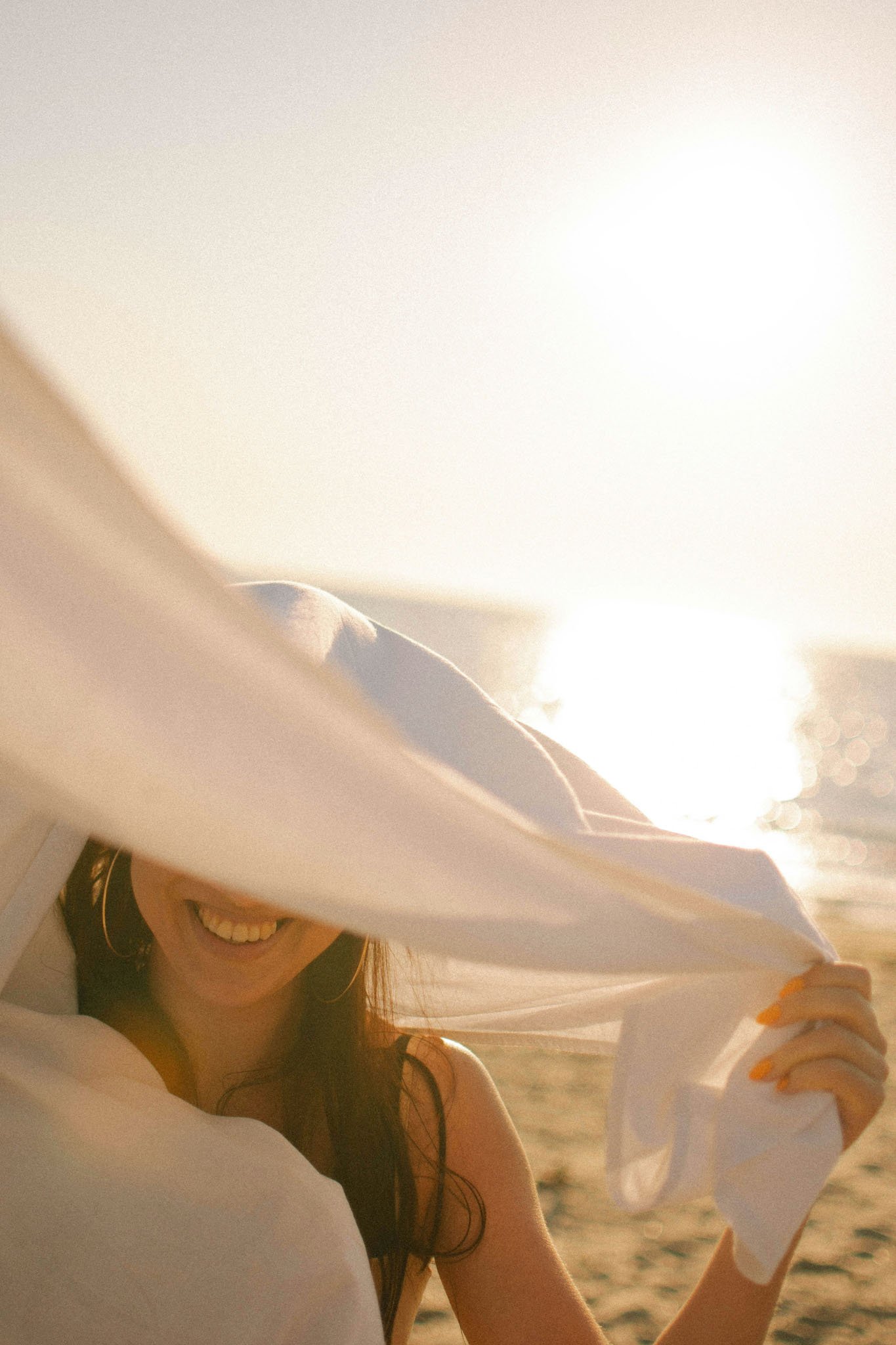 A smiling woman with long hair partially visible behind a person in the foreground, sitting on a beach near the ocean during sunset.