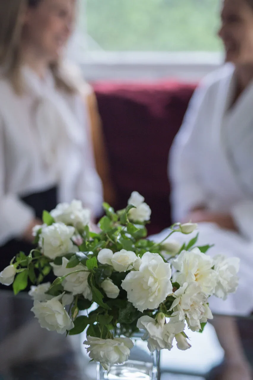 A bouquet of white roses and tulips in a clear glass vase on a table, with two women in white uniforms smiling and talking in the background.