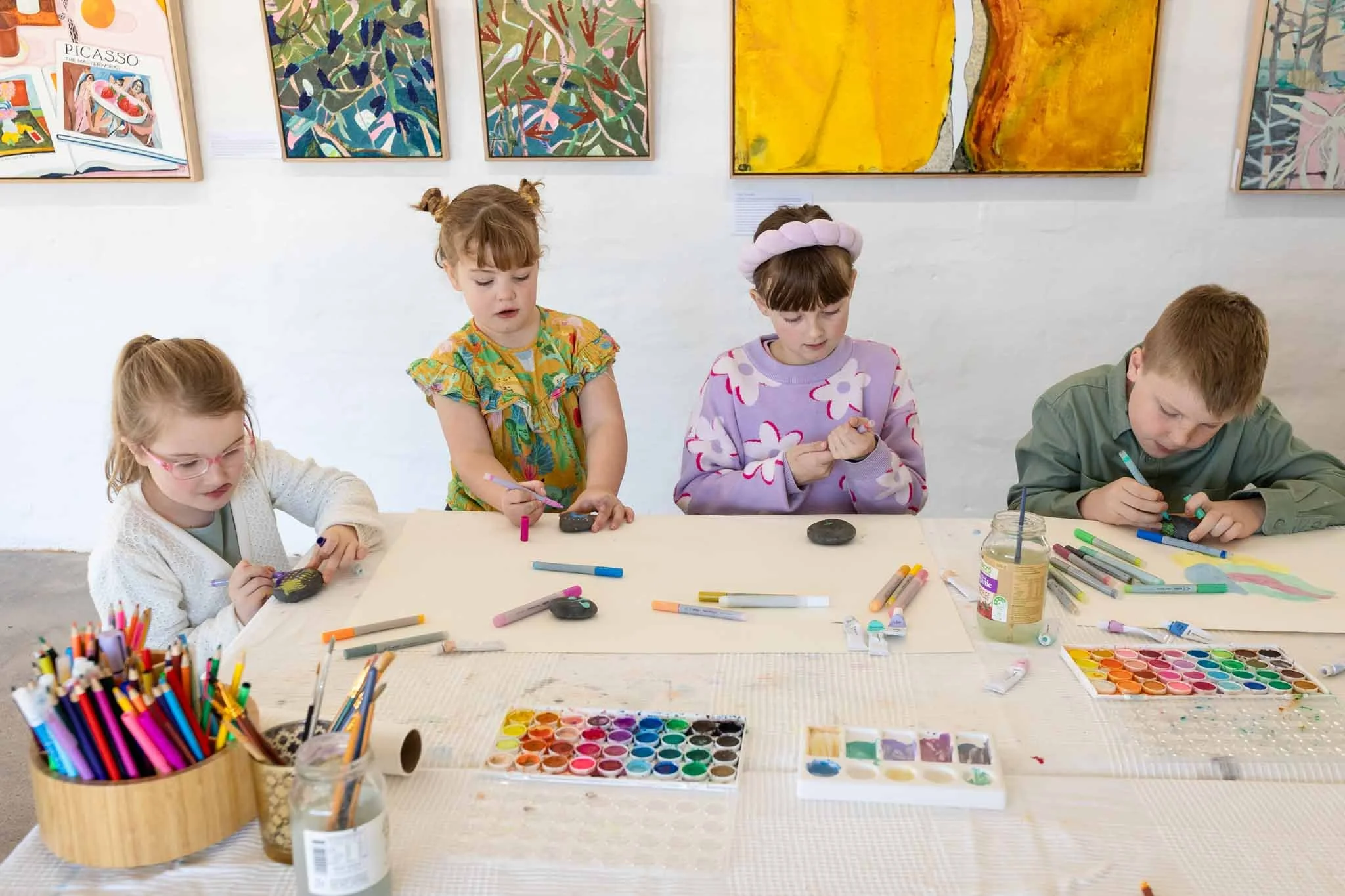 Children sitting at a table in an art gallery or classroom, painting and drawing with watercolors, colored markers, and stones, with colorful abstract paintings hanging on the wall behind them.