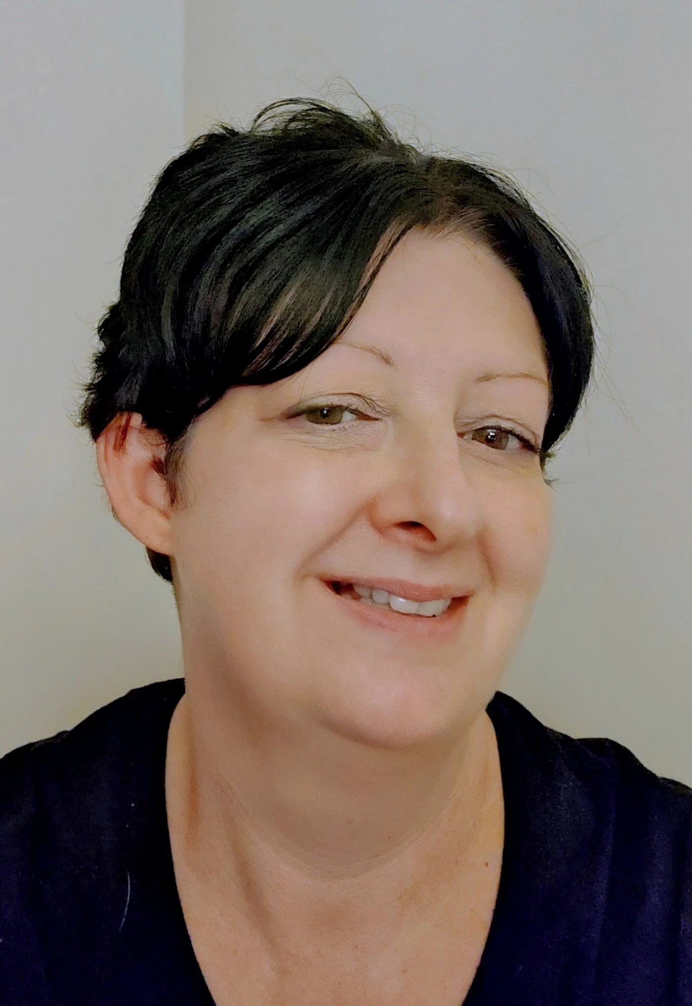 Close-up of a woman with short black hair, smiling, wearing a black top, against a plain light-colored background.