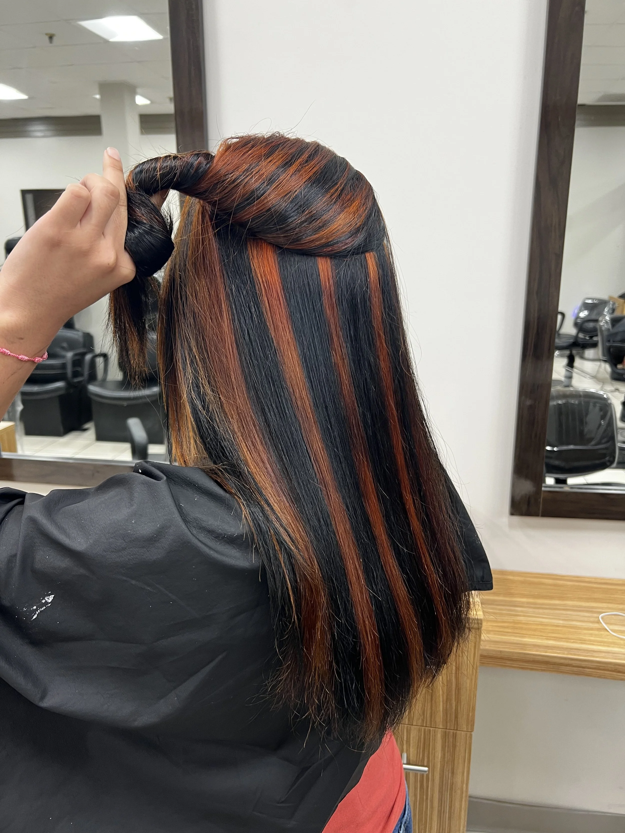 A person with long, straight hair decorated with orange and red highlights, sitting in a salon chair, while a stylist is working on the hair in a salon setting.