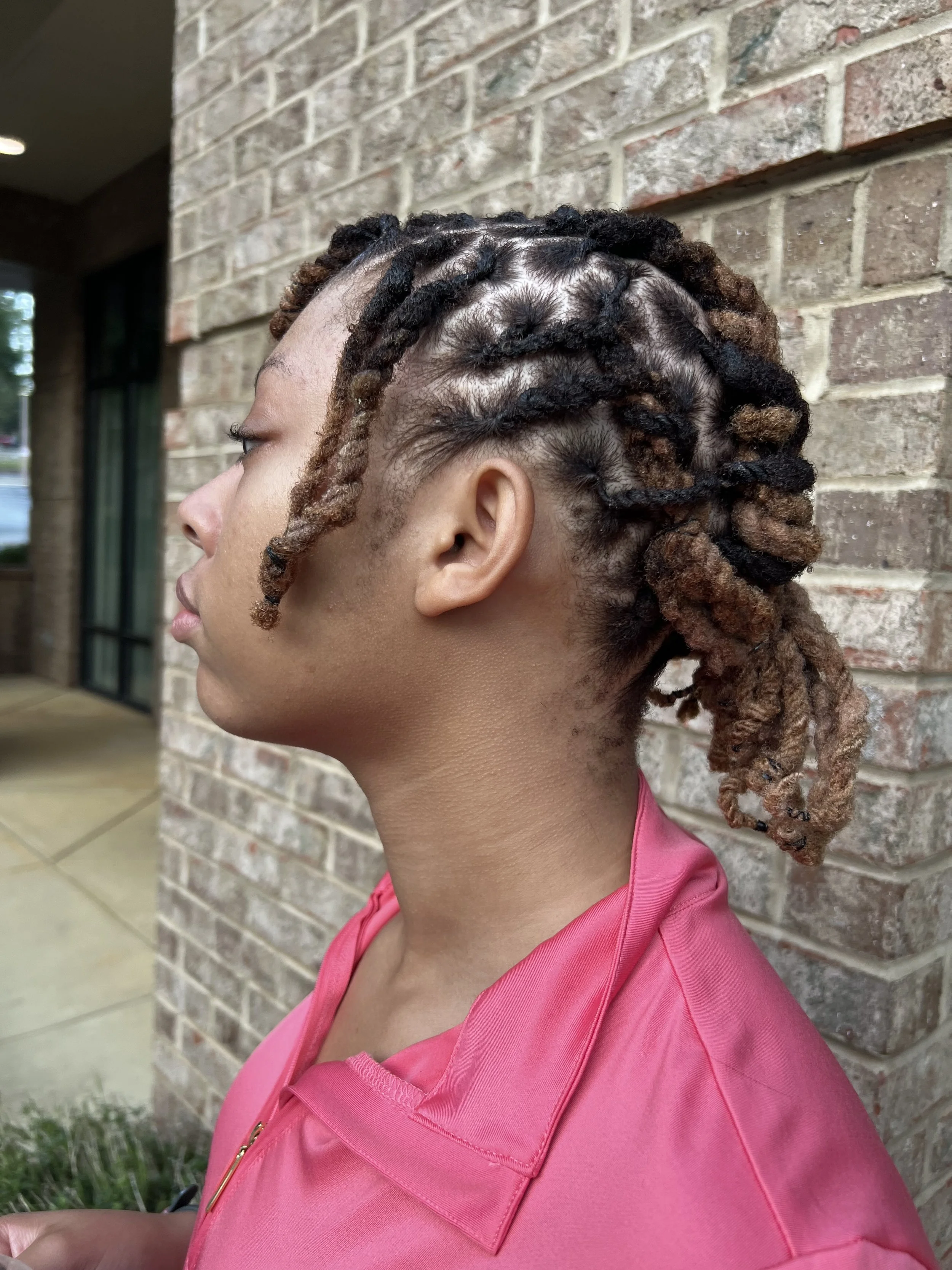 Profile of a woman with styled dreadlocks, wearing a pink jacket, standing against a brick wall.