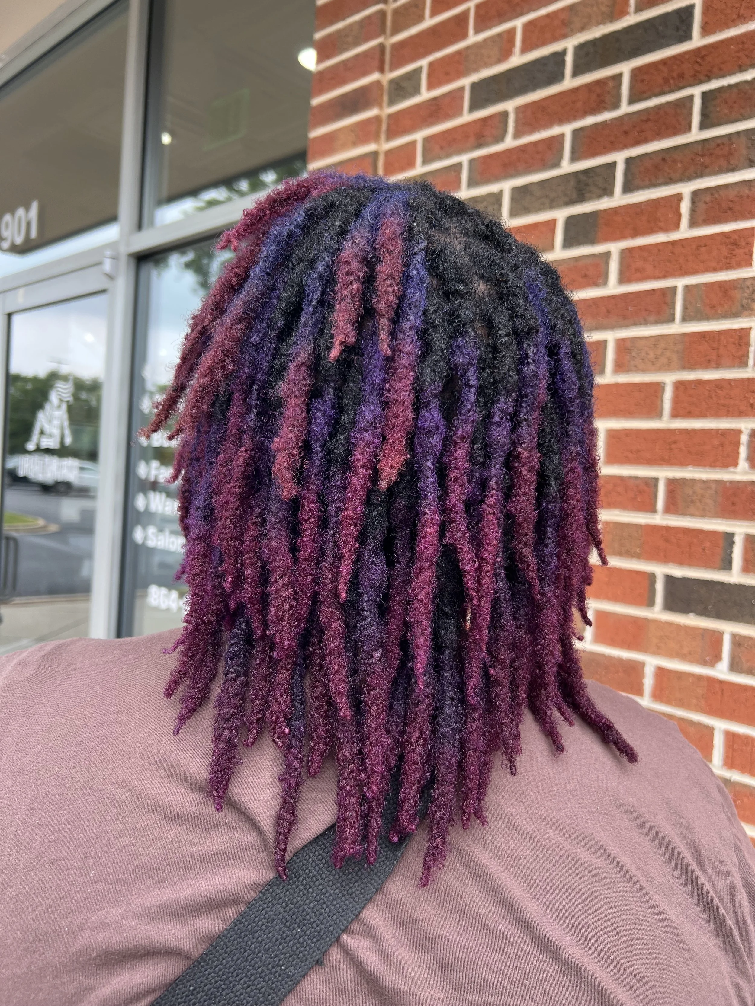 Close-up of a person's head with purple, black, and pink dreadlocks, sitting outdoors against a brick wall.