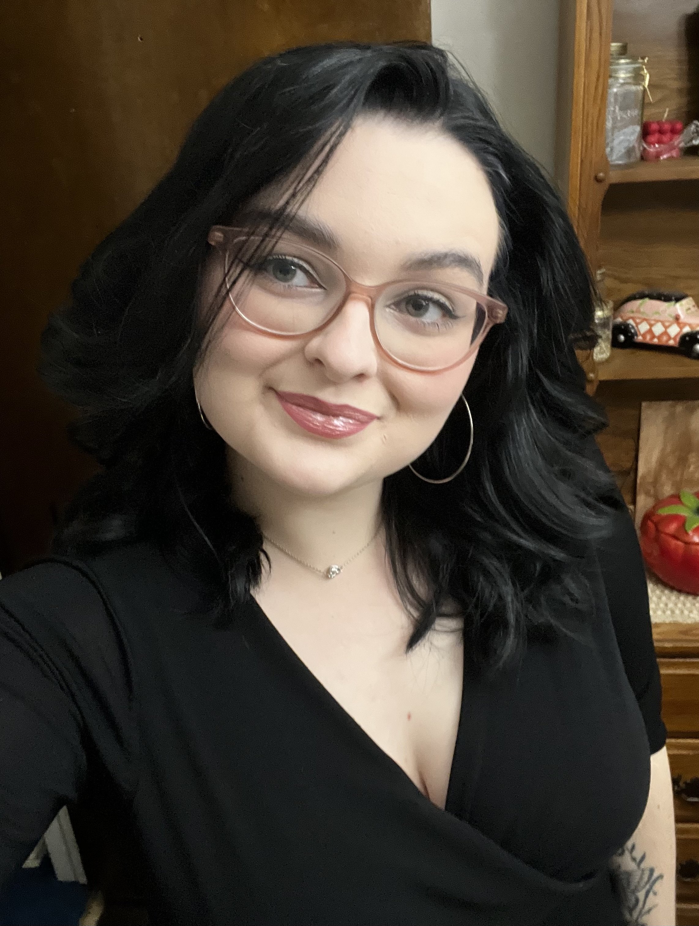 A woman with long black hair, wearing glasses and a black top, smiling in front of a wooden shelf with jars and decorations.