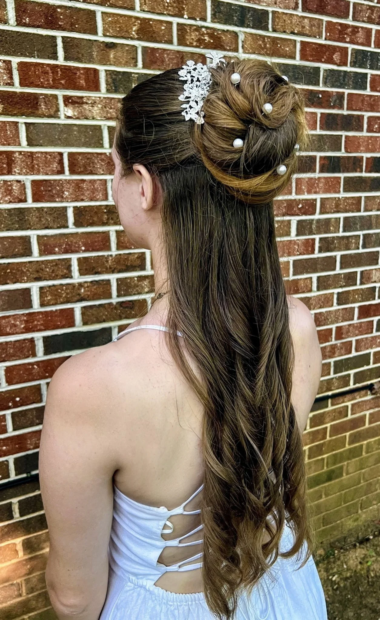 A woman in a white dress with lace-up sides, styled in a long, wavy hairstyle adorned with a pearl and floral hairpiece, standing against a brick wall.