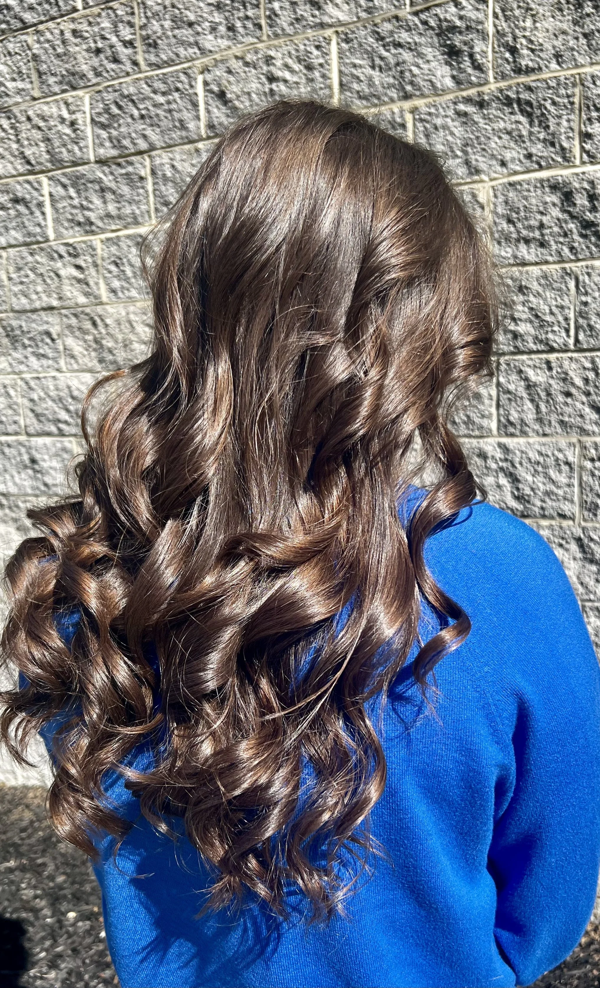 The image shows a person with long, brown, curly hair standing against a brick wall. The person is wearing a bright blue shirt.