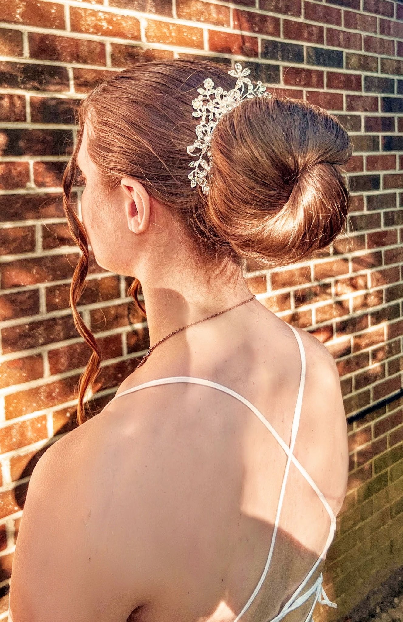 A woman with red hair styled in an elegant bun, wearing a decorative tiara, and a white dress with thin straps, standing against a brick wall.