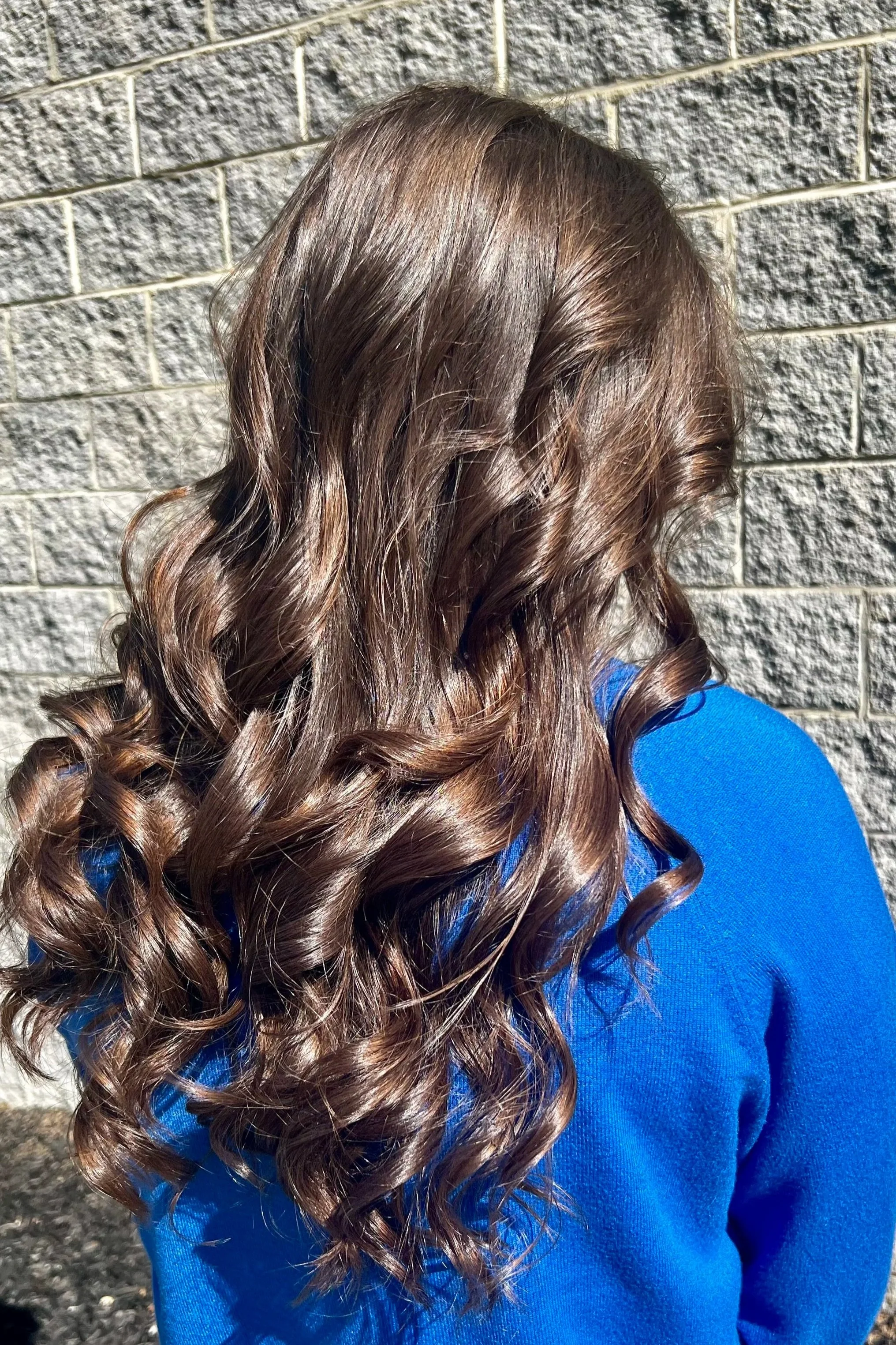 Close-up of a person's long, curly, brown hair with highlights, wearing a royal blue top, standing against a textured stone wall.