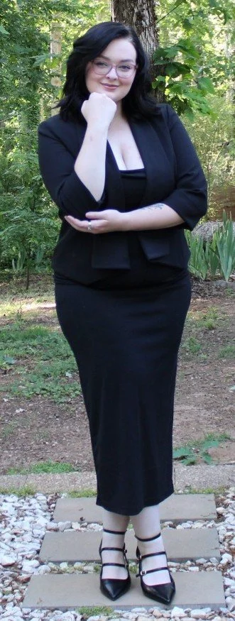 Woman with black hair and glasses standing outdoors on a pathway, wearing a black dress, blazer, and heels, surrounded by greenery.