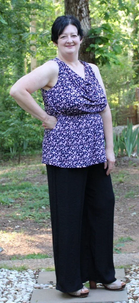 Woman standing outdoors in a garden, smiling, wearing a sleeveless purple floral top and black pants.