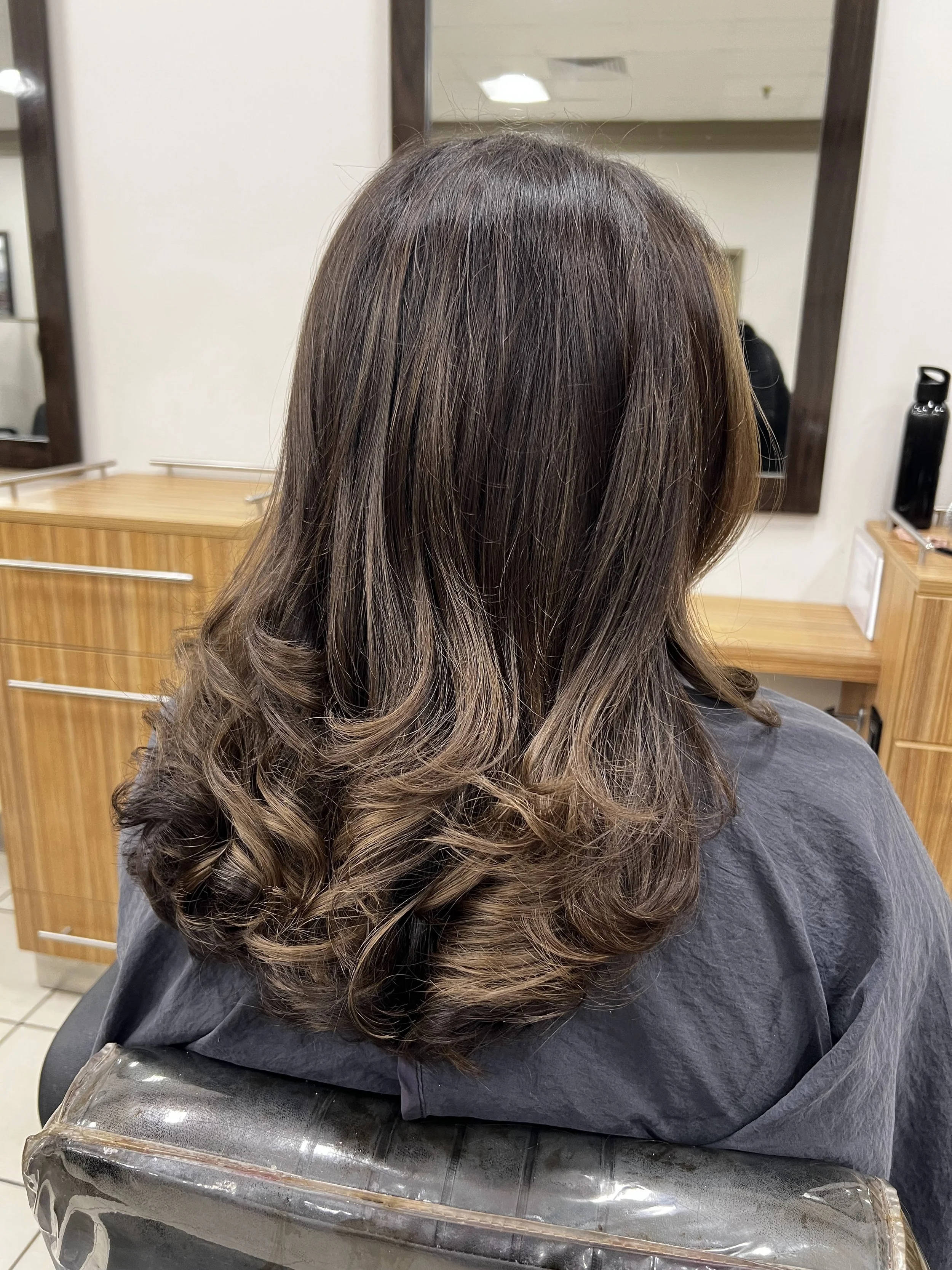 Woman with long, wavy, brown hair sitting in a salon chair, facing away from the camera.