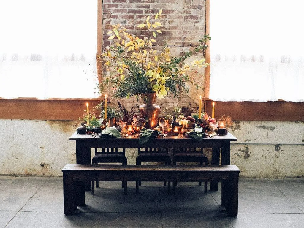 Decorative table setting with candles, flowers, and foliage in a rustic indoor space.