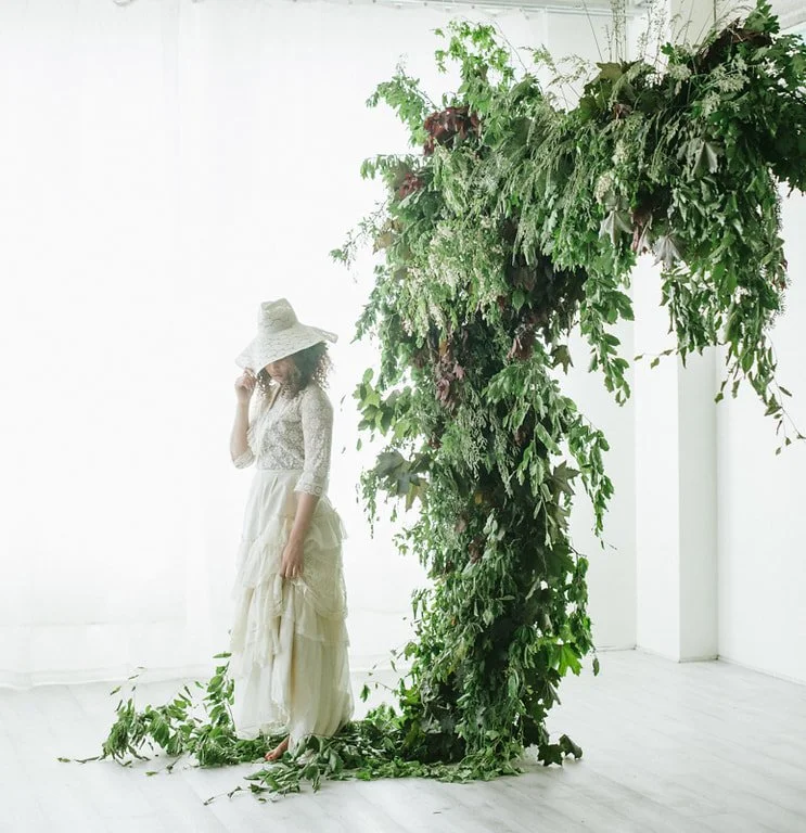 A woman dressed in a white lace blouse and flowing skirt, wearing a wide-brimmed hat, stands next to a large, lush green plant formation shaped into a letter or figure, with some foliage on the floor.