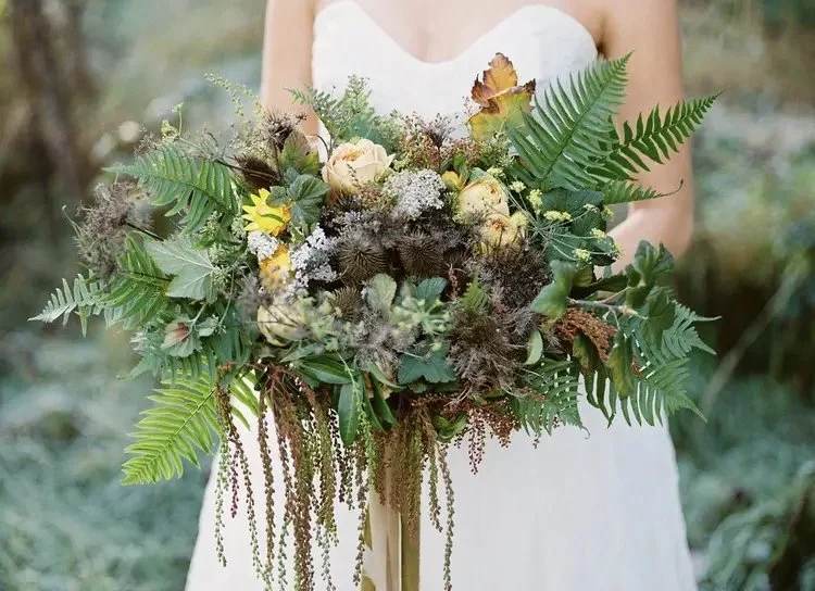 A bride holding a large bouquet of flowers and greenery outdoors.