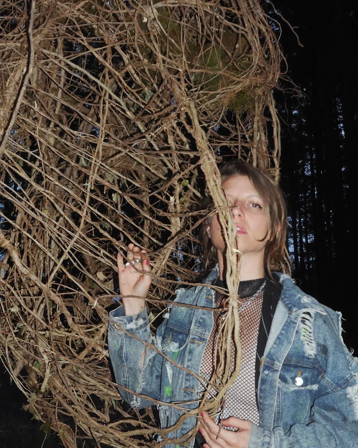 A young woman with short, wavy hair, wearing a distressed denim jacket over a black fishnet top, is standing in a dark forest, holding a large tangled pile of dry branches and vines, partially obscuring her face.
