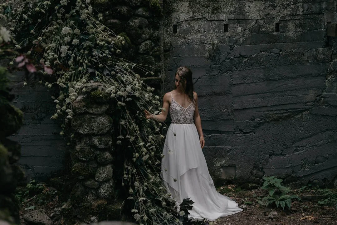 A woman in a white wedding dress standing next to a stone wall and a stone column with cascading flowers and greenery.