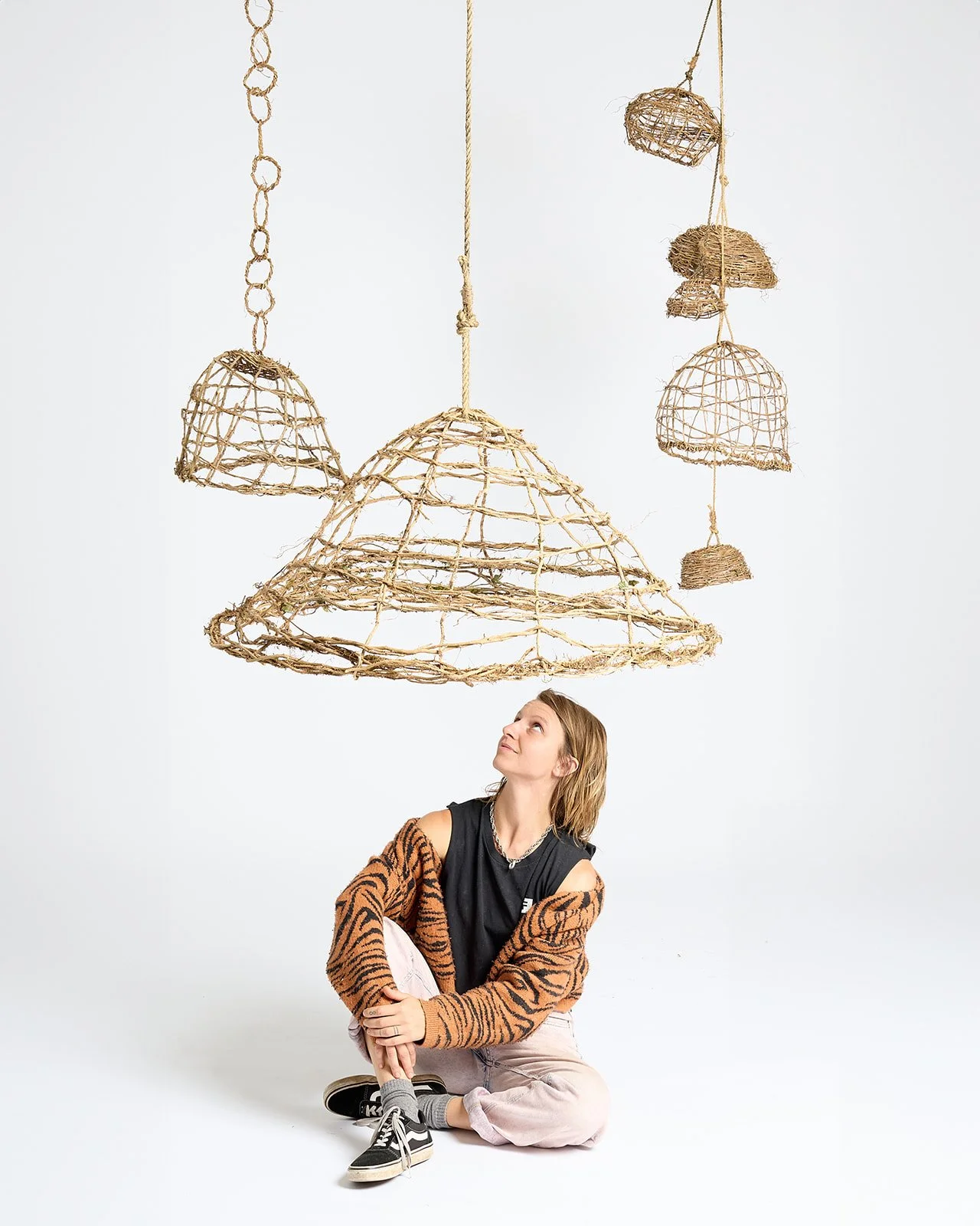 A young woman with blonde hair, wearing a tiger-striped jacket, white pants, and black sneakers, is sitting on the floor looking up at hanging wicker lamps.