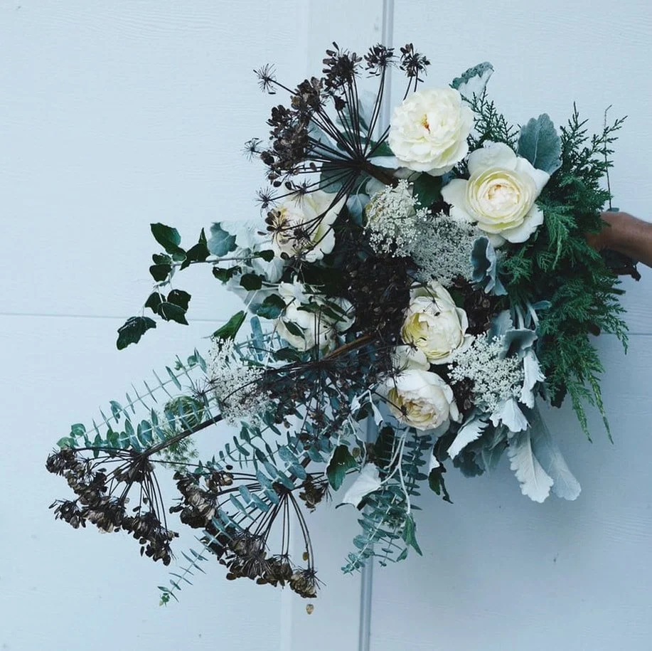 A bouquet of white roses, baby's breath, and various greenery, including ferns and eucalyptus, arranged against a light-colored background.