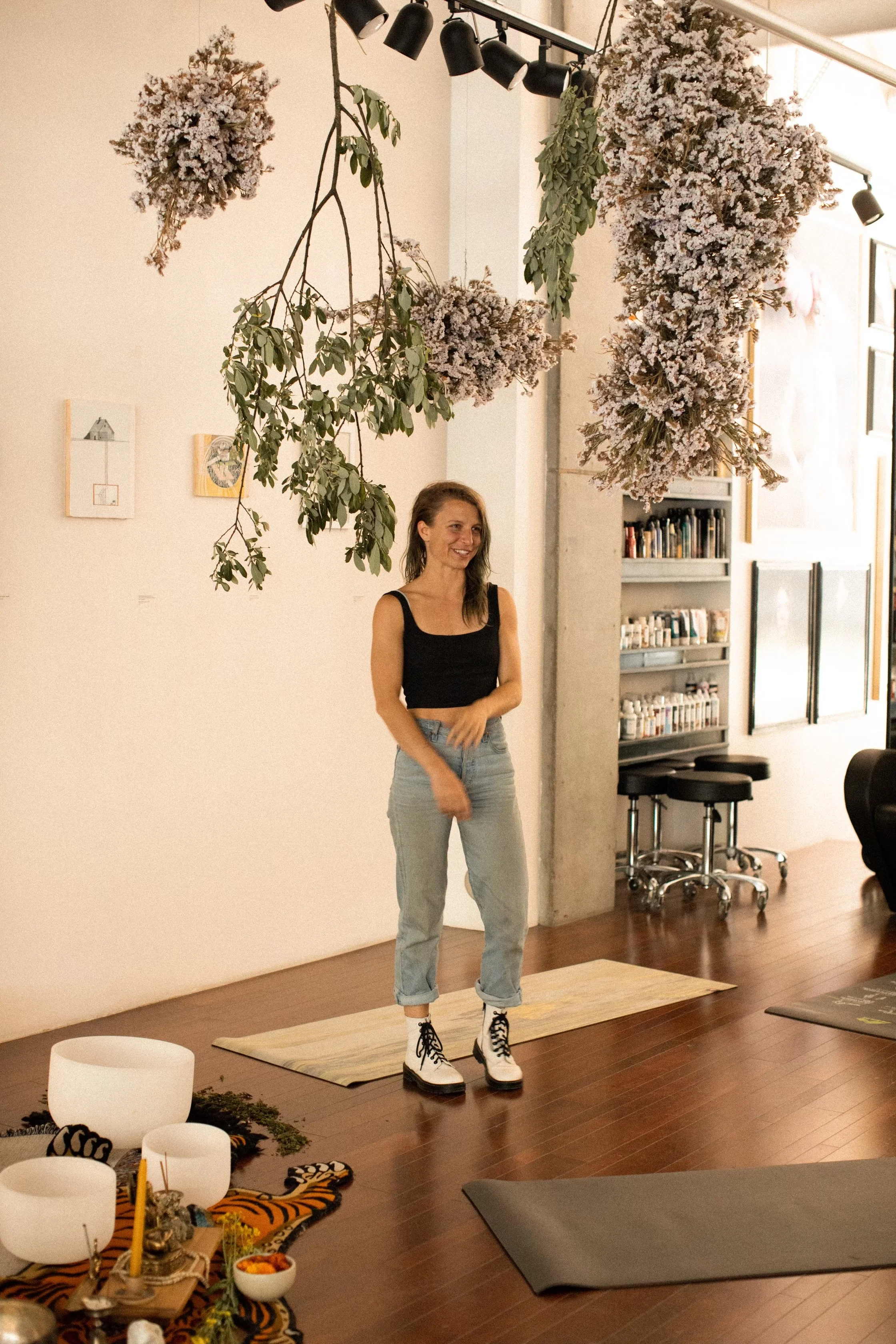 A woman stands on a yoga mat in a room decorated with hanging flowers, artwork, and a shelf with books, smiling.