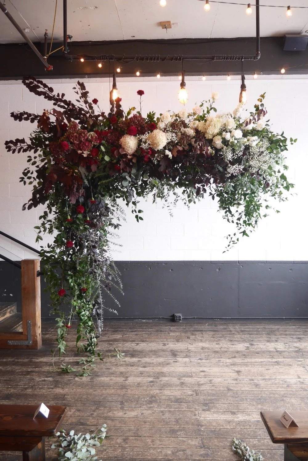 A large floral arrangement in an indoor space with string lights overhead, featuring white, red, and dark purple flowers and green foliage, hanging above a wooden floor.