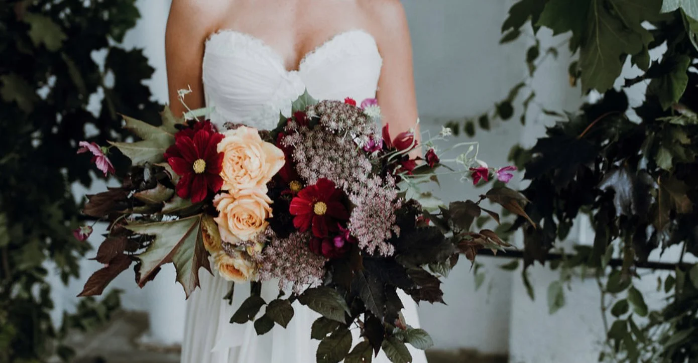 A woman in a white strapless dress holding a large bouquet of various flowers, including red, peach, and pink blooms, with dark green leaves around her.