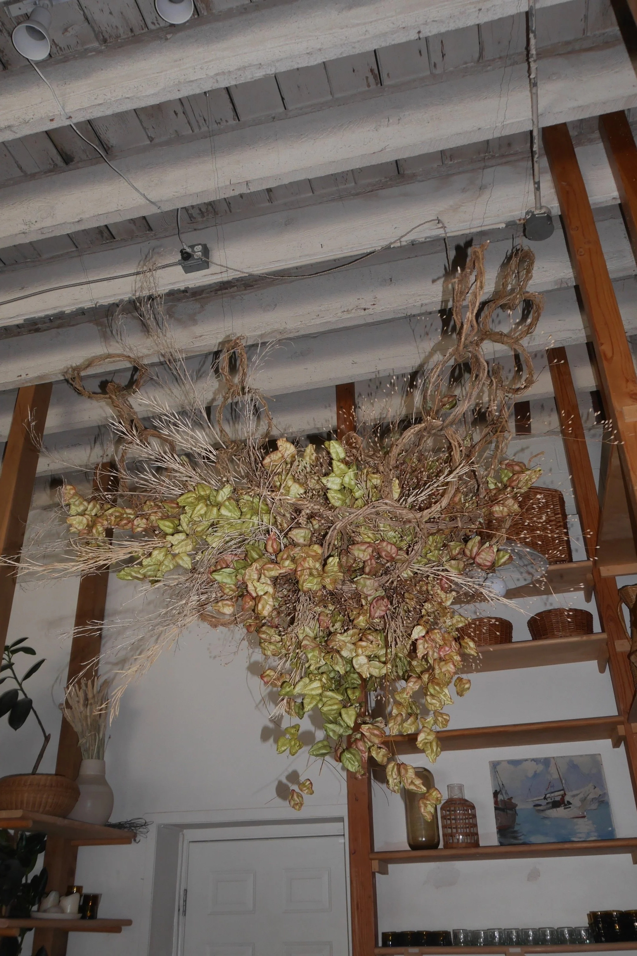 A large dried floral arrangement with twigs, leaves, and flowers hangs from a wooden ceiling beam in a cozy interior space. The ceiling has exposed beams and electrical wiring. There are decorative items on wooden shelves and a painting on the wall.