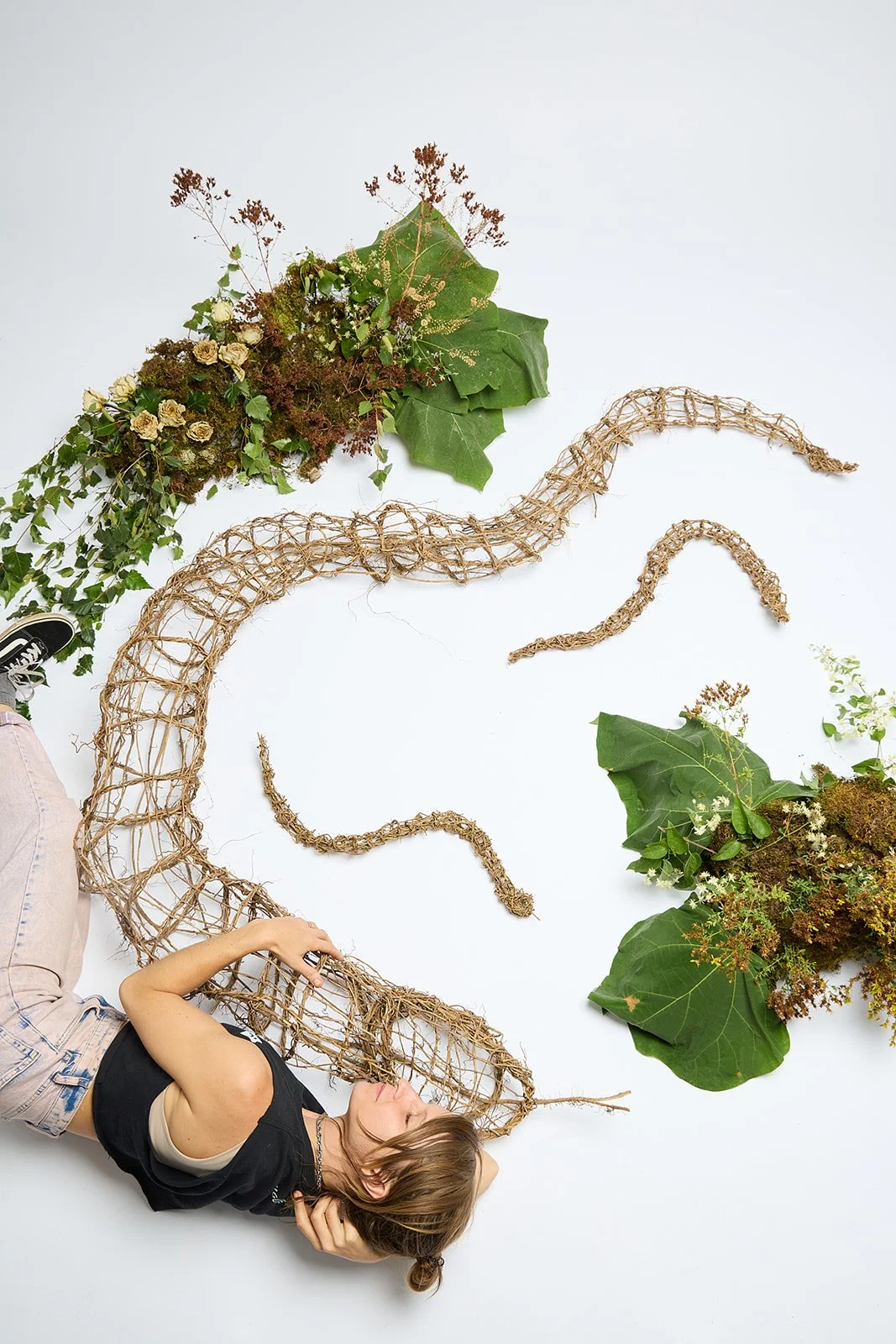 A woman lying on the floor with her head resting on her hand, surrounded by large green leaves, moss, flowers, and a long twisted straw or wicker sculpture.