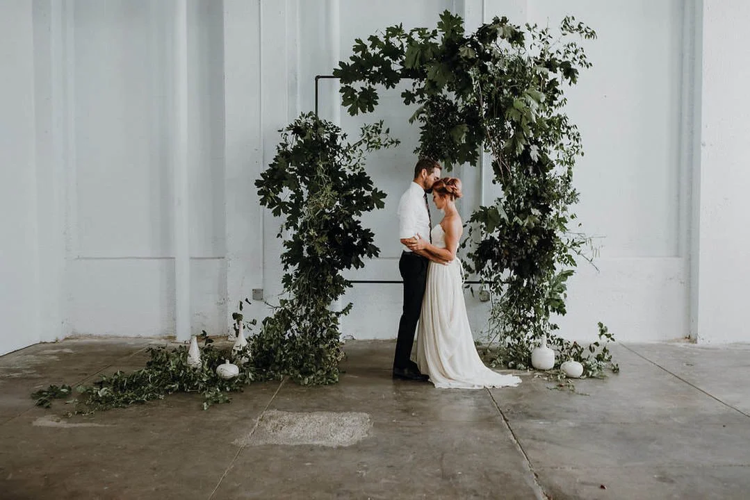 A couple dressed in wedding attire standing close together under a green leafy floral arch in an industrial-style venue with white walls and concrete floor.