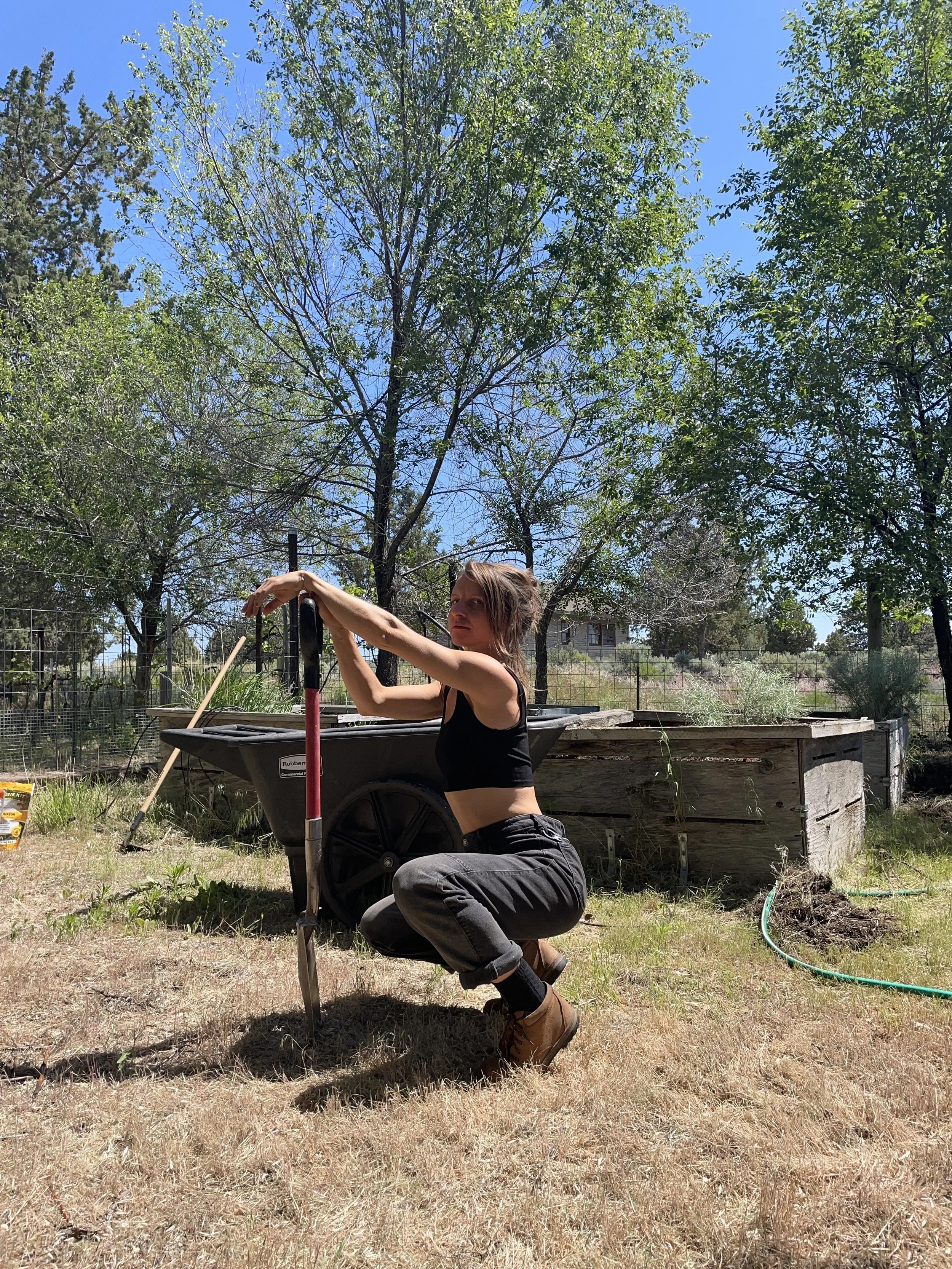A young woman with short brown hair kneeling on the ground outdoors in a yard with trees and a wooden raised garden bed. She is wearing a black sleeveless crop top, dark pants, and tan boots, and is holding a gardening tool. The sky is clear and blue