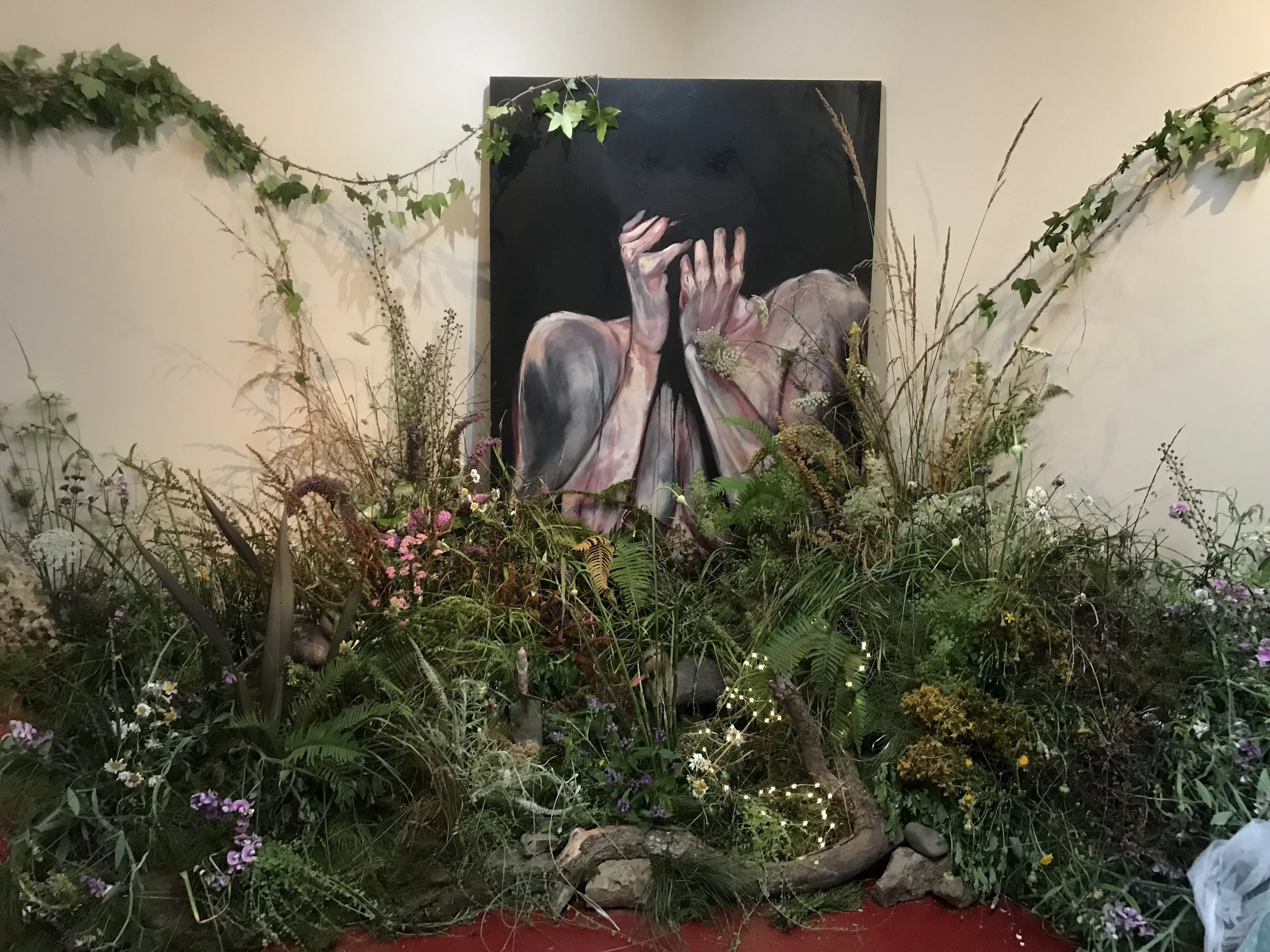 A painting of a person with short dark hair covering their face with their hands, surrounded by a large arrangement of various flowers and plants in front of a plain light-colored wall.