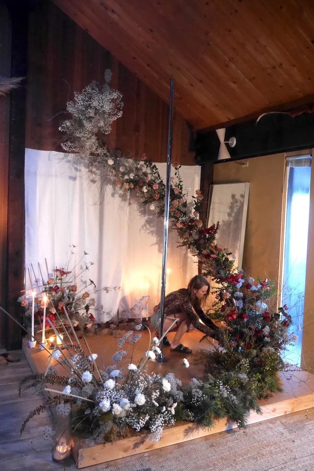 A woman arranging a floral display with an arch of flowers and greenery, surrounded by candles and additional flower arrangements in a decorated indoor setting.