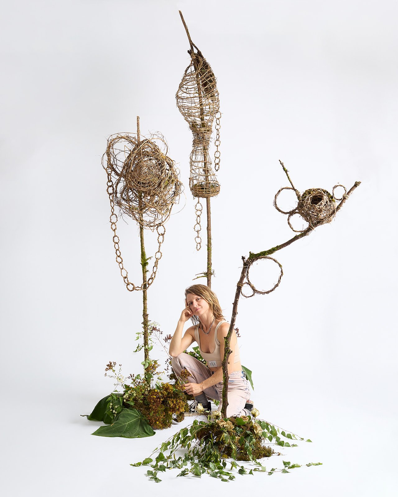 A woman with short hair sitting among two tall, thin structures made of intertwined twigs and branches, decorated with woven baskets and chains, with greenery at the base.