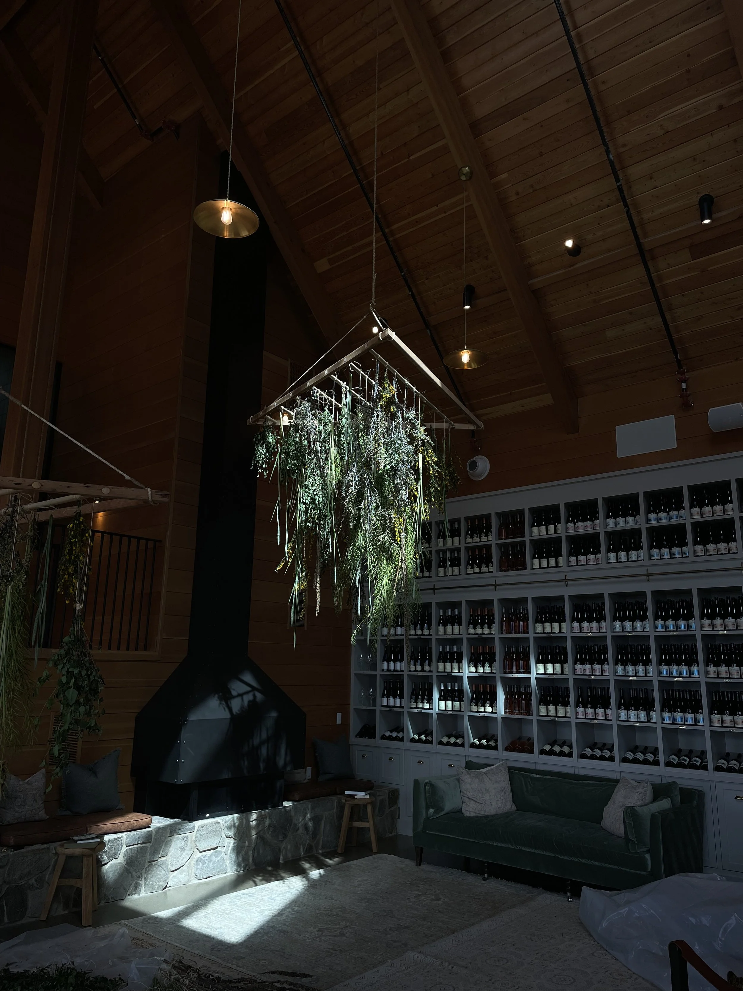 Living room with wooden ceiling, a black fireplace, hanging plants, a dark sofa with pillows, and a large storage unit filled with bottles or jars, illuminated by sunlight and ceiling lights.