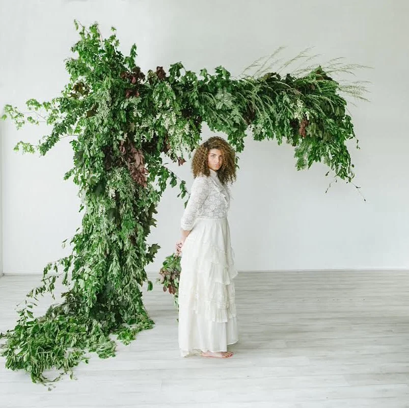 A woman standing in a minimalistic room with a large, lush, curved green plant arching over her.
