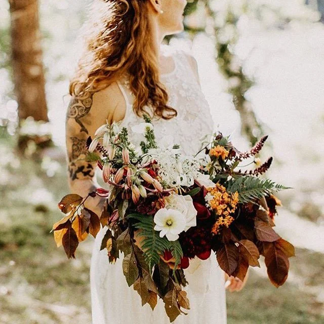 A woman with long, curly red hair, wearing a white lace dress, holding a large bouquet of assorted flowers and autumn leaves outdoors.