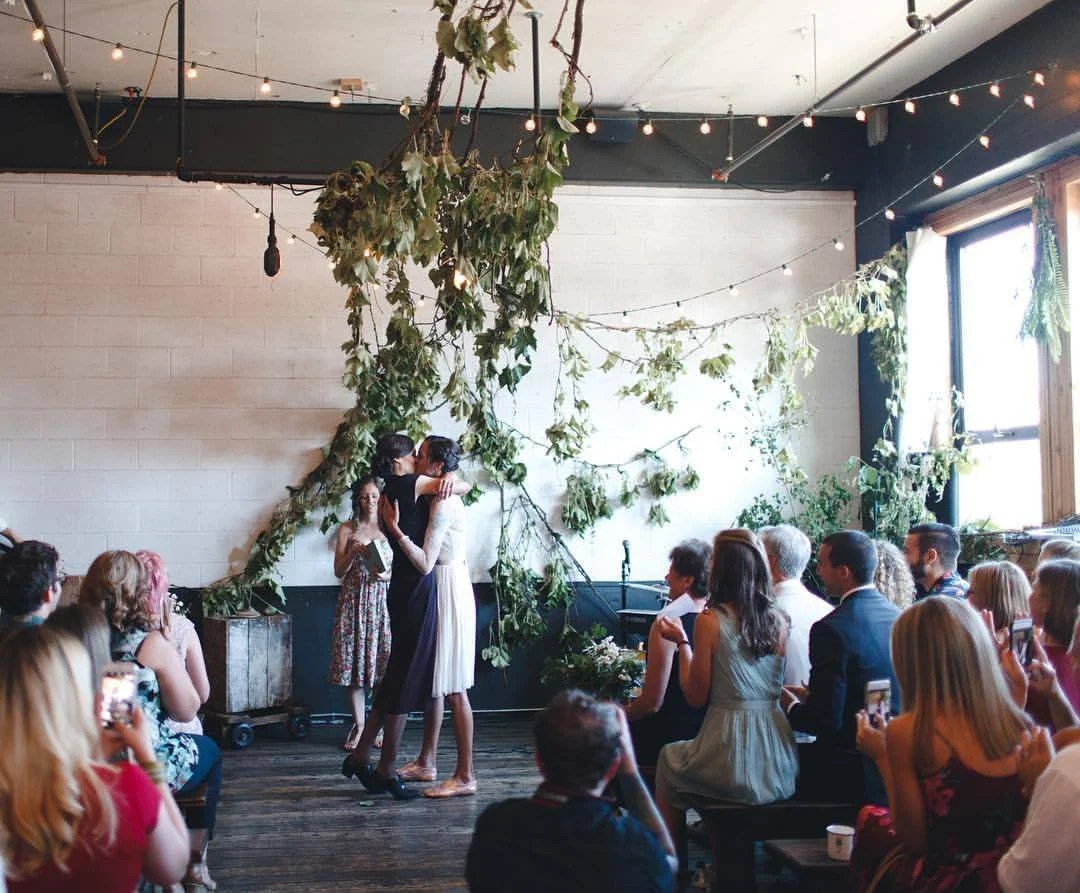 A wedding ceremony with two women hugging and kissing, surrounded by seated guests in a decorated indoor space with string lights and greenery.