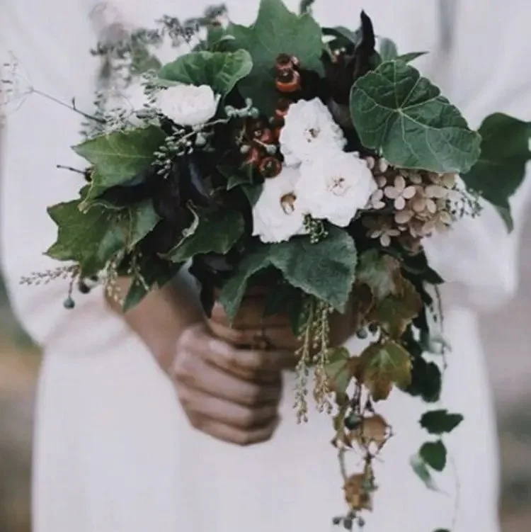 Person holding a bouquet of white flowers, green leaves, and small decorative elements.