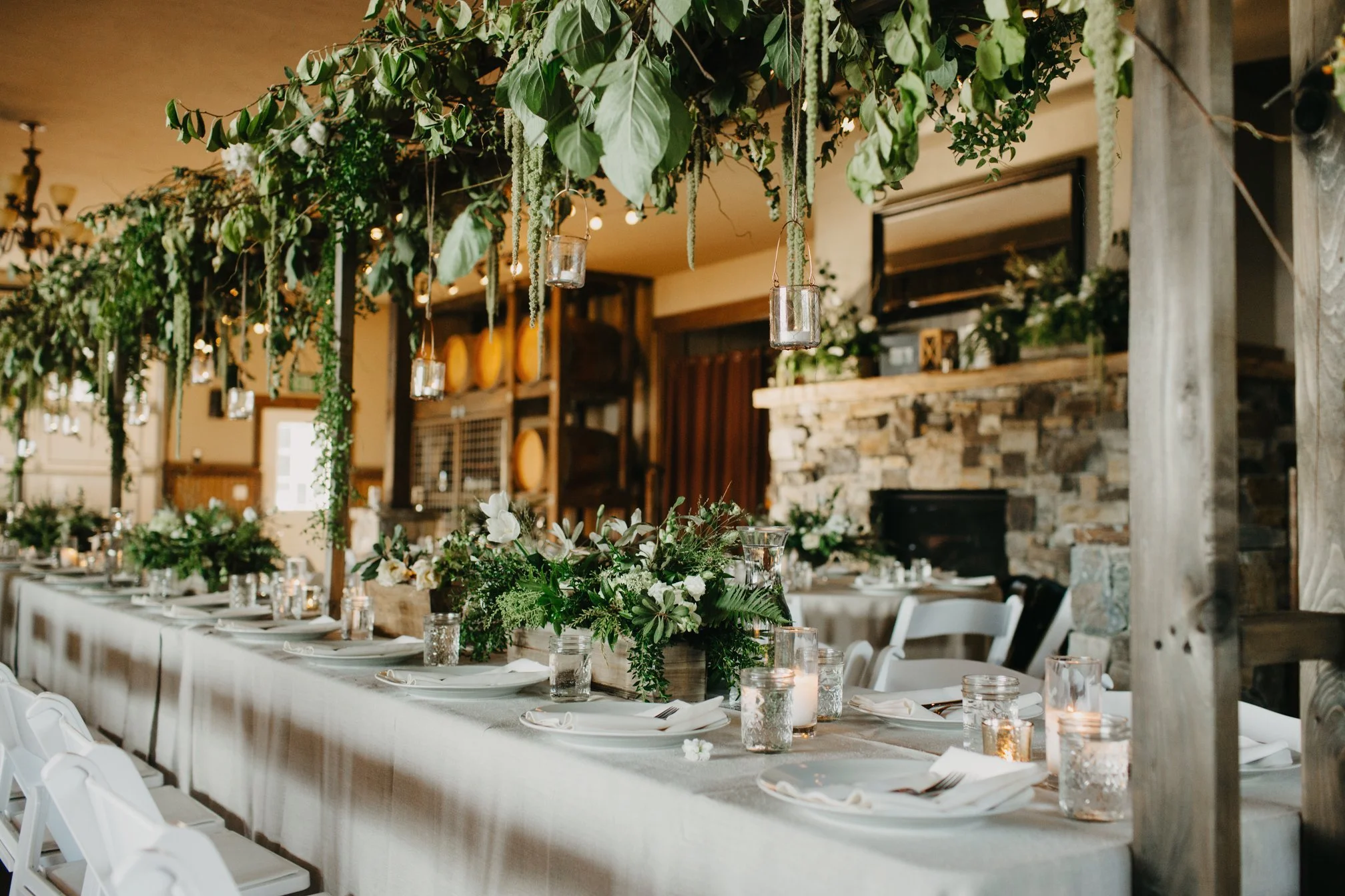 A decorated dining hall with a long table adorned with floral arrangements, candles, glassware, and neatly placed plates and silverware, under a hanging greenery and candle decorations, in a rustic setting with stone and wood accents.