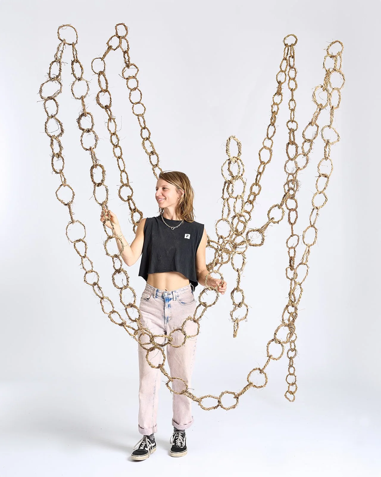 A young woman in a black sleeveless top and light jeans is standing inside a large heart-shaped outline made of thick, twisted rope links, laughing and looking to her left.