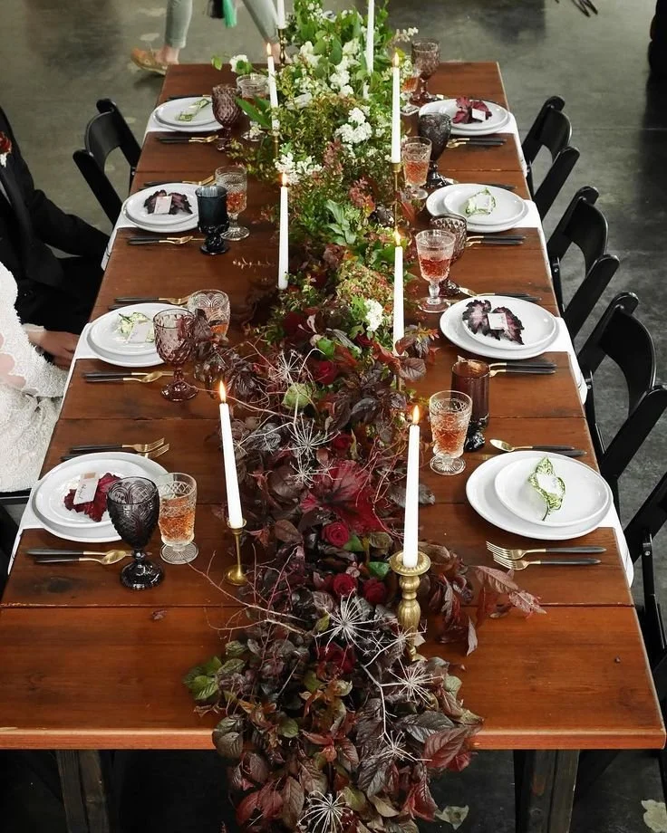 A long wooden dining table decorated with a central floral and foliage arrangement, candles, and arranged tableware for a formal meal, set in an indoor venue.