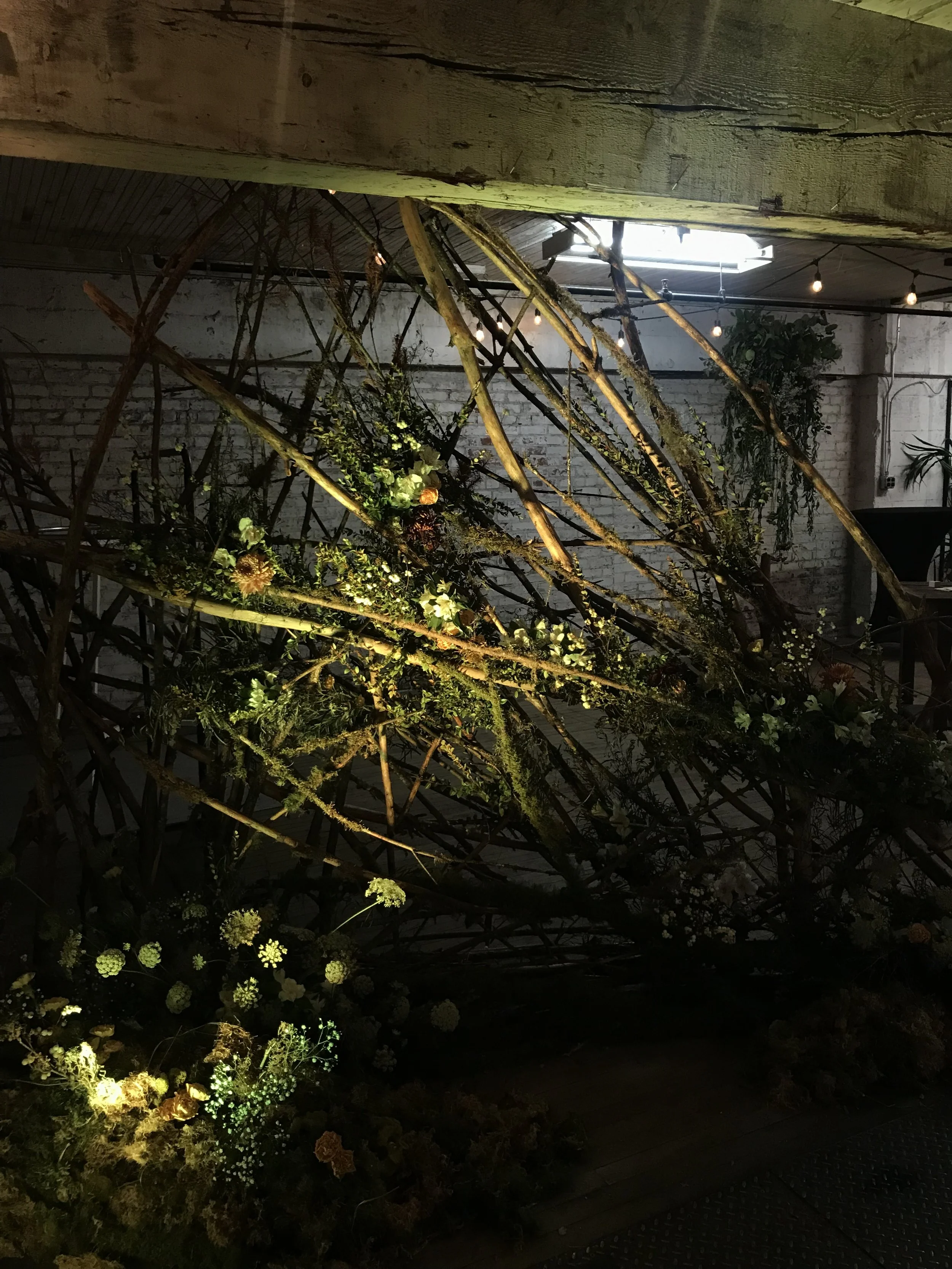 Decorative installation with intertwined branches and flowers, illuminated by soft lighting in an industrial-style space with exposed brick walls and a skylight.