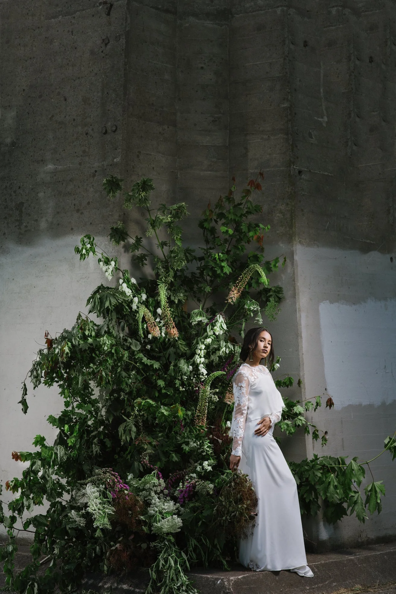A woman in a white bridal gown standing against a large concrete wall with green and white flowers and plants behind her.