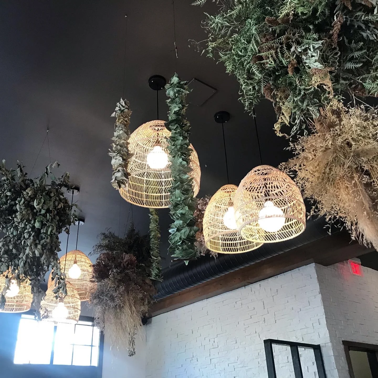 Interior view of a modern space with hanging wicker light fixtures, ceiling greenery, and a white brick wall.