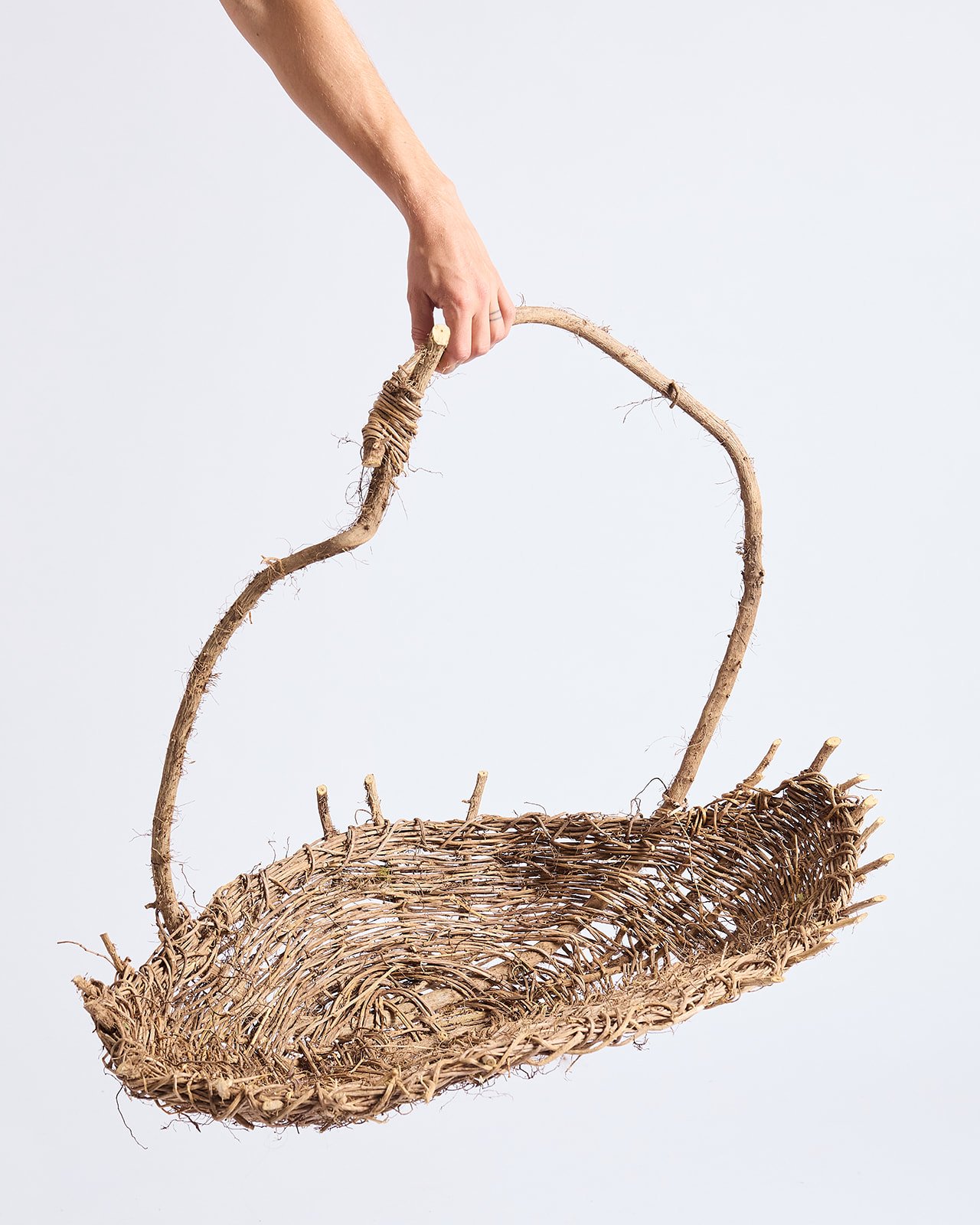 A hand holding the handle of an empty woven basket made of natural materials, against a plain background.