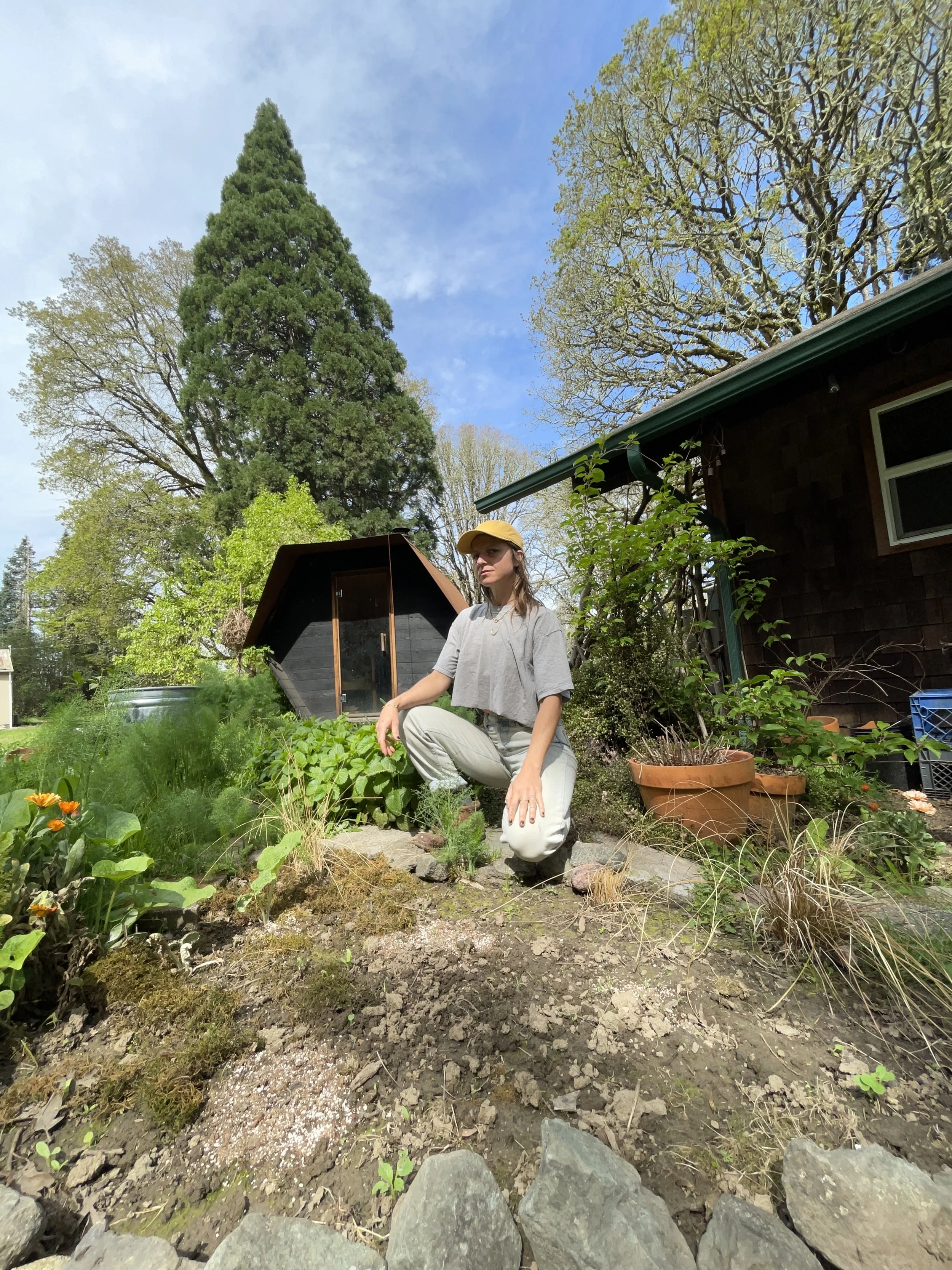 A woman wearing a yellow hat and gray shirt kneeling in a garden with various plants, a house with a brown roof, and tall trees in the background on a sunny day.