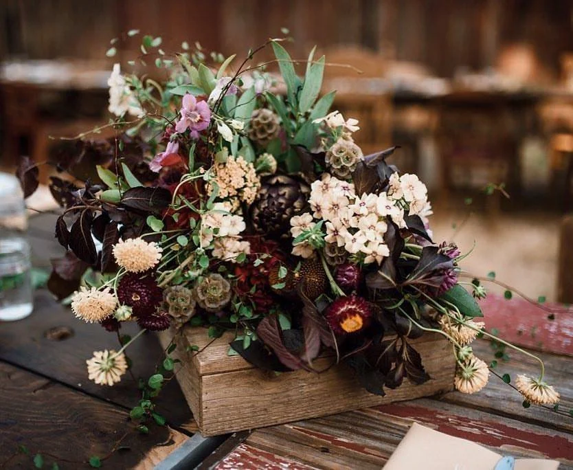 A floral arrangement with mixed flowers and greenery in a wooden box on a rustic table.