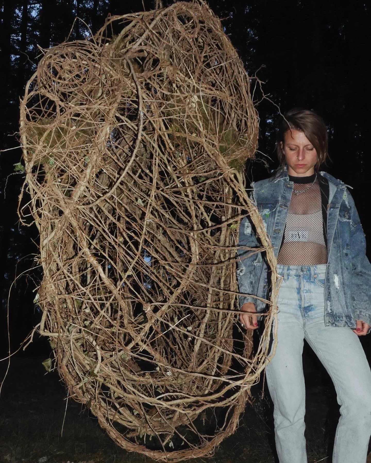 A young woman with short brown hair, wearing a denim jacket, a black fishnet top, white jeans, and a nose ring, standing outdoors at dusk with a large, abstract, spherical sculpture made of intertwined dried twigs or vines.